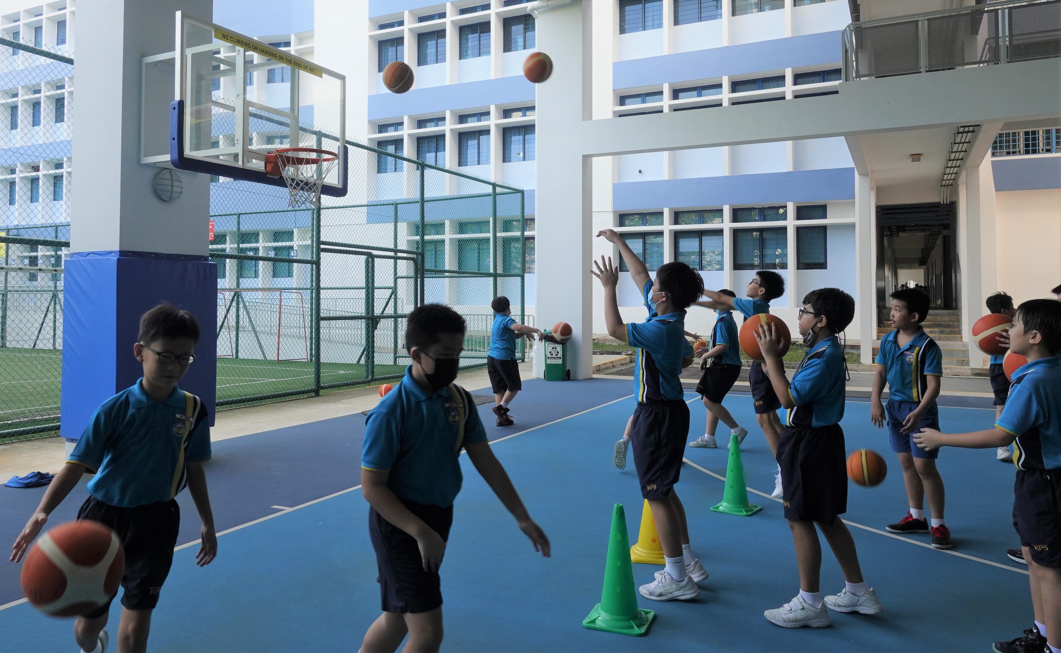 School children in blue uniforms practice basketball on an outdoor court with a hoop and green cones.